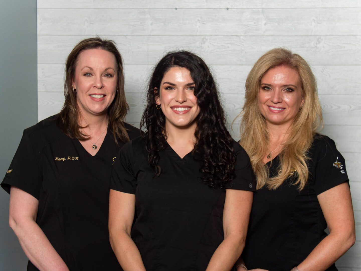 Three women in black outfits smiling against a plain wall.